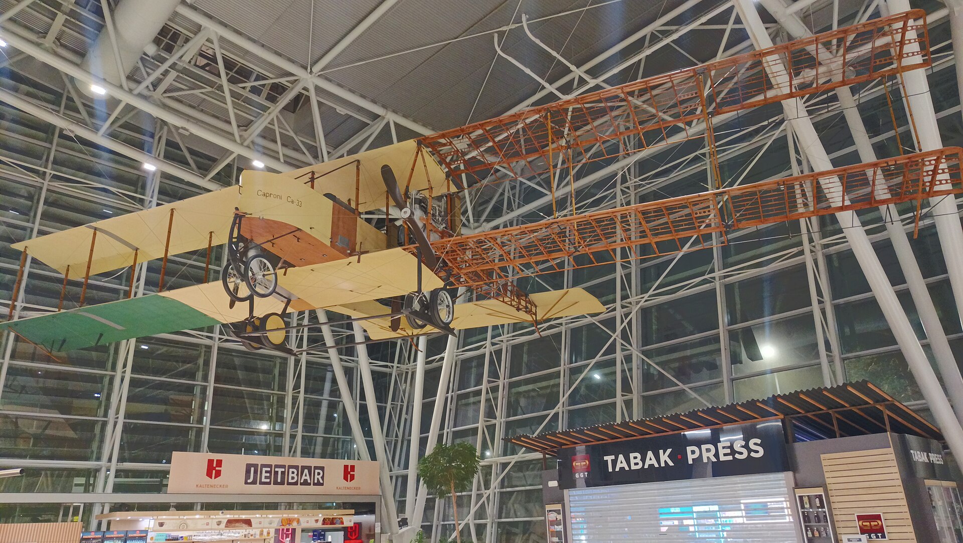 Historické lietadlo Caproni Ca-33 zavesené pod stropom odletovej haly letiska, pod ním novinový stánok a slnečník kaviarne