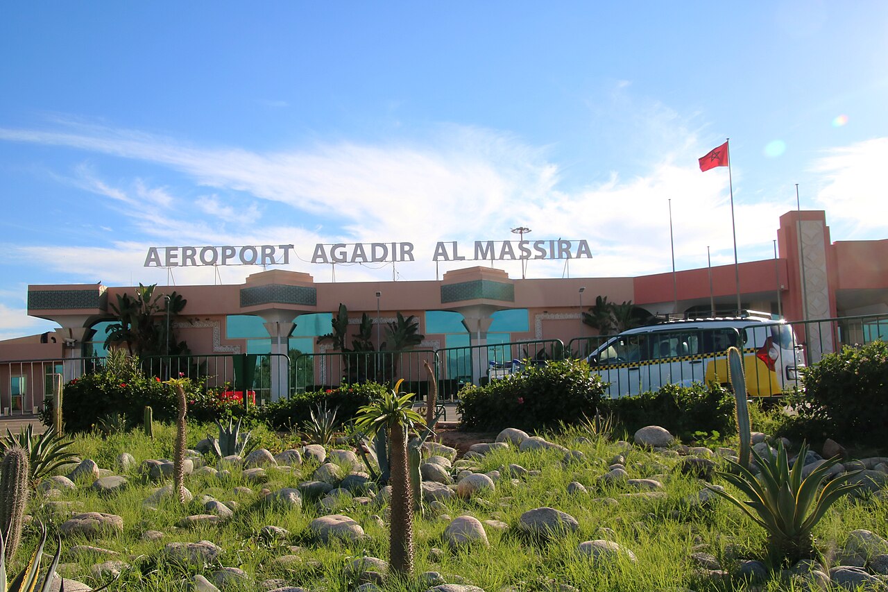 Nízka budova terminálu letiska Agadir – Al Massira s veľkým nápisom na streche, pred ňou zeleň a modrá obloha